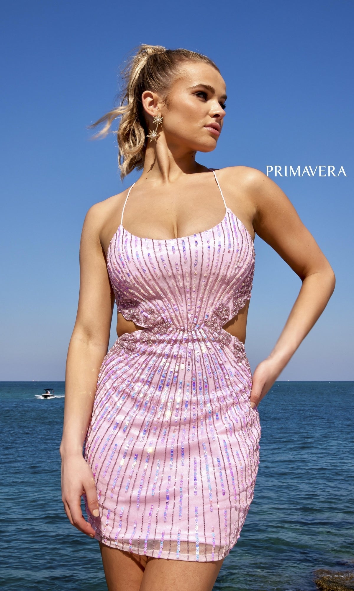 A woman wears the Short Sequin Homecoming Dress with Cut Outs 4025, a strappy pink dress with an open back, standing by the ocean under a clear blue sky.